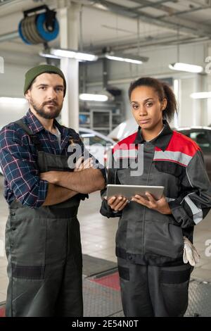 Zwei junge ernsthafte Kollegen in Uniform stehen vor Kamera, während einer von ihnen mit Tablet in zeitgenössischen Workshop Umwelt Stockfoto