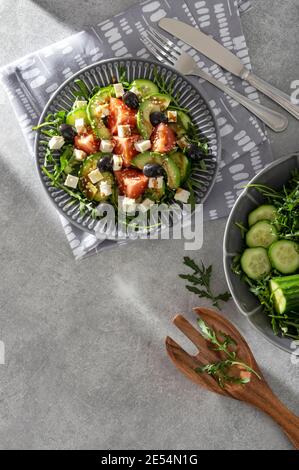 Frischer vegetarischer Salat aus Rucola, Gurke, Tomaten, Käse, Avocado und Oliven, garniert mit Balsamico-Glasur und Sesam auf grauem Stein Stockfoto