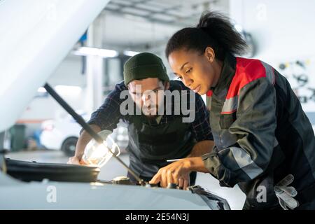 Junger bärtiger Mann in Arbeitskleidung, der Lampe hält und sich umbeugt Öffnen Sie die Haube, während seine Kollegin auf den Automotor zeigt Während der Arbeit Stockfoto