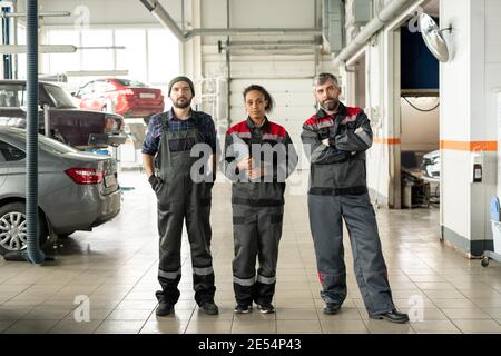 Drei seriöse interkulturelle Profis in Arbeitskleidung stehen vor Kamera in großer Werkstattumgebung am Ende der Arbeit Tag Stockfoto