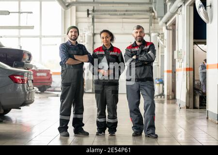 Junge fröhliche interkulturelle Arbeiter des Auto-Wartungszentrums stehen in Große Werkstatt vor der Kamera am Ende von Arbeitstag Stockfoto