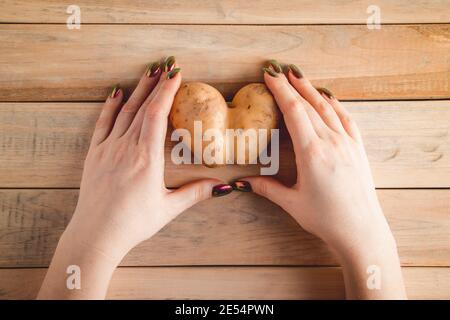 Herzförmige Kartoffeln in den Händen auf Holzhintergrund. Valentinstag-Konzept. Hässliches Gemüse. Stockfoto