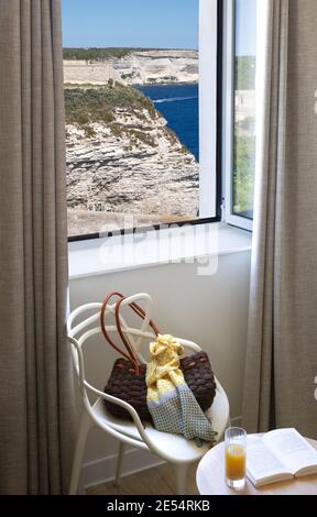 Frühstück auf einem Tisch und weißem Stuhl mit Tasche und Schal, Fenster mit Blick auf das Meer und die Klippen Stockfoto