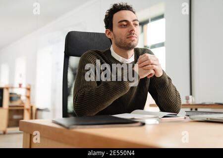 Lächelnder, selbstbewusster junger Unternehmer, der am Schreibtisch sitzt Stockfoto