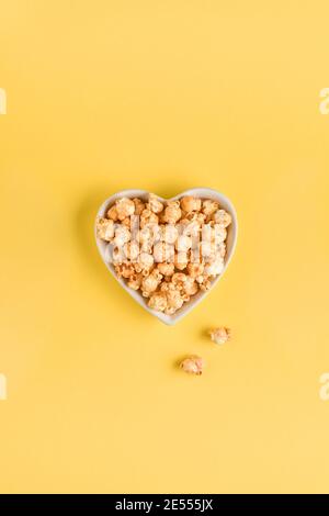 Köstliches süßes Popcorn mit Karamell in weißem Keramik-Herzteller, isoliert auf Trendfarbe gelber Hintergrund. Stockfoto