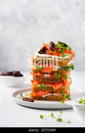 Sandwiches stapeln sich mit geräucherten rosa Lachsoliven, kalamata, Microgreens und Frischkäse auf grauer Keramikplatte und trendigem Betonhintergrund. Traditio Stockfoto