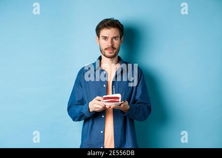 Bild von lächelndem jungen Mann, der Geburtstag feiert, den B-Day-Kuchen mit brennender Kerze hält und auf die Kamera schaut, auf blauem Hintergrund stehend Stockfoto