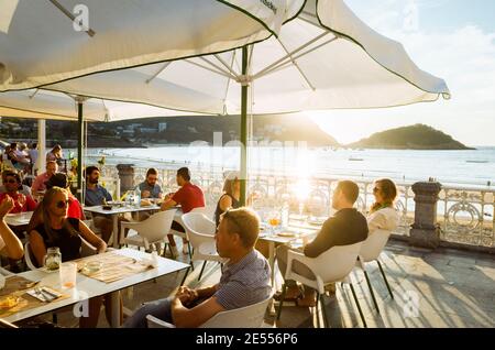 Donostia, Gipuzkoa, Baskenland, Spanien - 15. Juli 2019: Die Menschen entspannen bei Sonnenuntergang in einem schicken Straßencafé an der Strandpromenade von La Concha. Stockfoto
