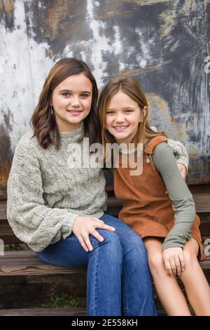 Zwei junge Schwestern sitzen nebeneinander und umarmen sich Mit den Armen umeinander im Winter sitzen In der Nähe einer städtischen Lagerhauswand Stockfoto