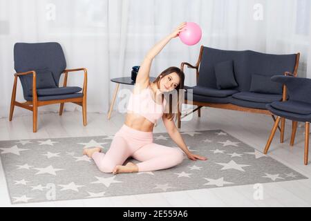 Fit Frau in rosa Trainingsanzug sitzt auf dem Boden und Macht seitwärts Kurven mit einem Ball in der Hand gestreckt Über ihrem Kopf nach Hause Stockfoto