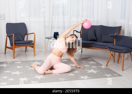 Schöne europäische Frau in rosa Trainingsanzug sitzt auf dem Boden Und macht seitwärts Kurven mit einem Ball in der Hand Gestreckt über ihren Kopf nach Hause Innenraum Stockfoto