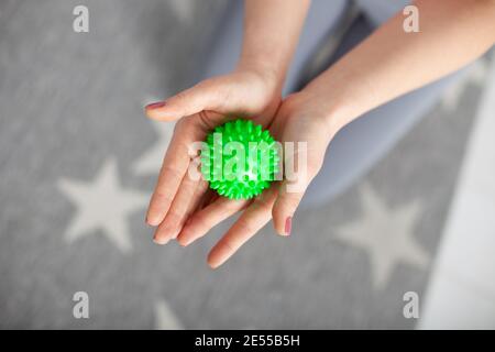 Nahaufnahme zwei Frauenhänden halten einen grünen Spikes Massage Ball Gegen einen grauen Teppich mit weißen Sternen und Knien Graue Leggings Stockfoto