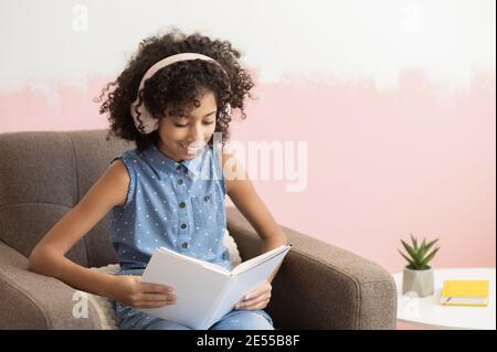 Ein kluger afrikanischer präteen Schulmädchen, das Freizeit mit verbringt Ein Buch Lesung sitzen am Sessel zu Hause Stockfoto
