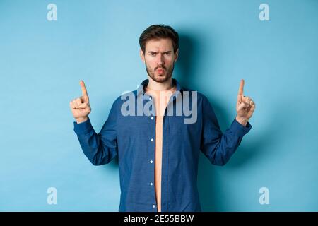 Skeptischer junger Mann runzelte die Stirn, zeigte die Finger nach oben und beschwerte sich, sah enttäuscht oder genervt aus, stand auf blauem Hintergrund Stockfoto