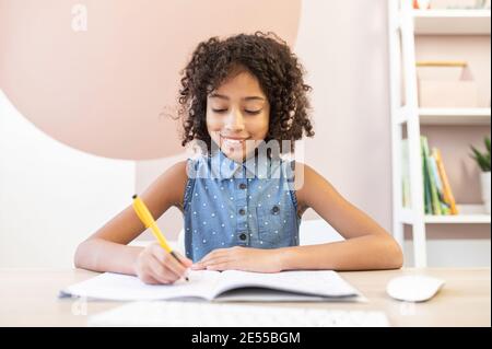 Ein Porträt von einem hübschen lockigen afrikanischen Schulmädchen trägt einen Kopfhörer, ein hübsches biracial Mädchen füllen Übungen Buch mit einem Stift, tun Hausarbeit Stockfoto