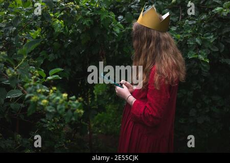 Präteen Mädchen trägt rotes Kleid und Papier Krone im Stehen Nass Garten mit Telefon Stockfoto