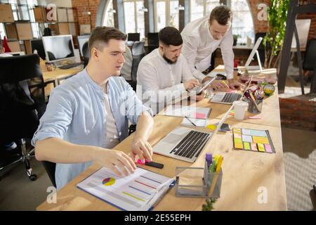 Erstellung von Jahresberichten. Junge kaukasische Kollegen arbeiten in einem Büro mit modernen Geräten und Gadgets zusammen. Schauen Sie beschäftigt, attened, fröhlich, erfolgreich. Konzept von Geschäft, Büro, Finanzen. Stockfoto