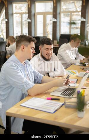 Erstellung von Jahresberichten. Junge kaukasische Kollegen arbeiten in einem Büro mit modernen Geräten und Gadgets zusammen. Schauen Sie beschäftigt, attened, fröhlich, erfolgreich. Konzept von Geschäft, Büro, Finanzen. Stockfoto