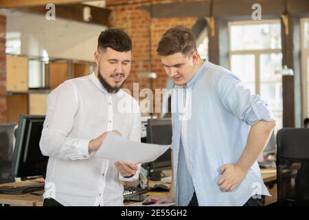 Erstellung von Jahresberichten. Junge kaukasische Kollegen arbeiten in einem Büro mit modernen Geräten und Gadgets zusammen. Schauen Sie beschäftigt, attened, fröhlich, erfolgreich. Konzept von Geschäft, Büro, Finanzen. Stockfoto
