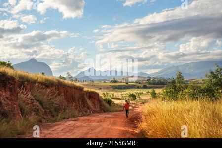 Ein einziger unbekannter junger madagassischer Mann mit Korb auf den Schultern, der über eine rote staubige Straße geht, nach Hause kommt, nachdem er die Felder bearbeitet hat Stockfoto