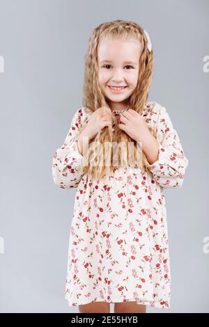 Porträt eines schönen lockigen kleinen Mädchens in einem eleganten Blumenkleid steht, lacht und schaut in die Kamera im Studio auf einem grauen Hintergrund Stockfoto