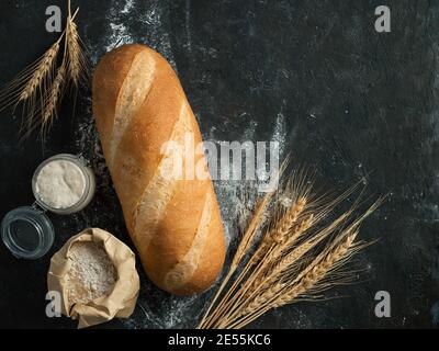 Britisches Weißblüher oder europäischer Sauerteig Baton-Brot auf schwarzem Hintergrund. Frisches Brot und Glas mit Sauerteig Starter, floer in Papiertüte und Ohren. Draufsicht. Kopierbereich Stockfoto