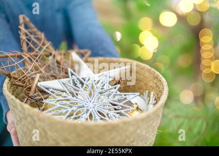 Die Hände des Kindes halten einen Korb aus Birkenrinde mit verschiedenen Weihnachtsdekorationen auf dem Hintergrund des hellen Bokeh auf dem Weihnachtsbaum. Stockfoto