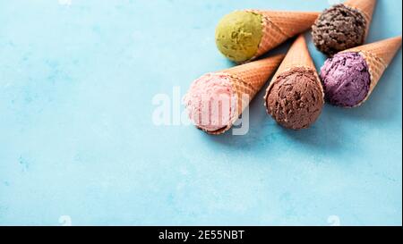 Verschiedene Eiscreme in Zapfen mit Kopierraum. Buntes Eis in Zapfen: Schokolade, Erdbeere, Heidelbeere, Pistazie oder Matcha-Tee, Kekse Schokoladenkekse Sandwich auf blauem Hintergrund. Banner Stockfoto