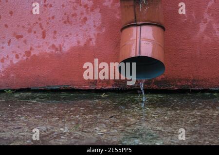 Ein dünnes Rinnsal Wasser fließt aus dem roten Metall Ablaufrohr auf dem Zement mit Kieselsteinen auf der eingestreut Blinder Bereich des Gebäudes mit dem roten b Stockfoto