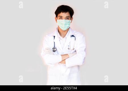 Ein Arzt, der mit gekreuzten Armen auf weißem Hintergrund posiert, trägt eine medizinische Gesichtsmaske und ein Stethoskop. Junge asiatische Arzt trägt einen weißen Mantel. Stockfoto