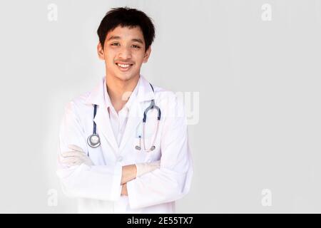Ein lächelnder Arzt, der mit gekreuzten Armen auf weißem Hintergrund posiert, trägt ein Stethoskop. Junge asiatische Arzt trägt einen weißen Mantel. Stockfoto