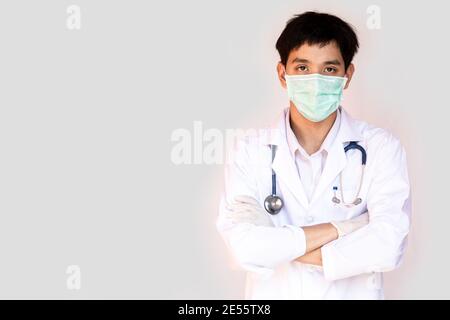 Ein Arzt, der mit gekreuzten Armen auf weißem Hintergrund posiert, trägt eine medizinische Gesichtsmaske und ein Stethoskop. Junge asiatische Arzt trägt einen weißen Mantel. Stockfoto