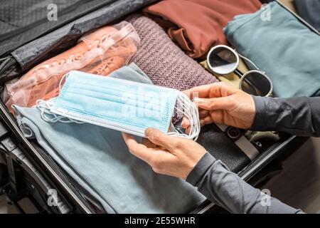 Eine Frauenhand steckt eine Maske in einen offenen Koffer, der mit Reisegepäck verpackt ist. Stockfoto