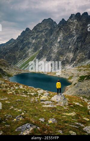 Wanderer Frau erkunden Sie die fantastische Landschaft und genießen Sie die Aussicht in der Nähe eines Bergsees in gelb in Jacke, hohe Tatra, Slowakei Stockfoto