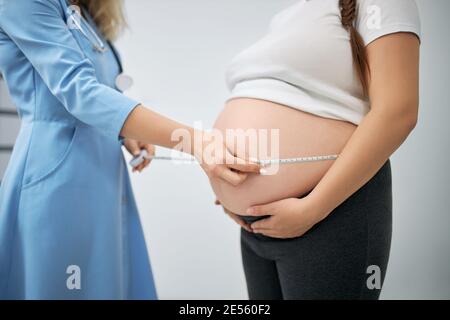 Qualifizierter Arzt, der den Bauch einer schwangeren Frau mit Klebeband in der modernen Klinik misst. Frauenärztin untersucht Wachstum des Fötus. Konzept der Gesundheitsversorgung. Stockfoto