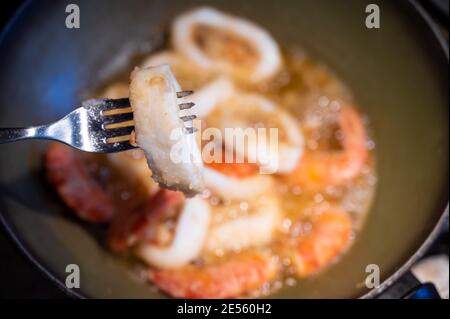 Häusliche Szene: Im Hintergrund bemehlte Garnelen und Tintenfischringe in kochendem Öl gebraten. Im Vordergrund ein Tintenfisch-Ring an der Spitze einer Gabel Stockfoto