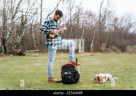 Hispanischer Mann spielt ein Lied auf seiner Ukulele in der Wald mit seinem Hund auf dem Gras liegen Stockfoto