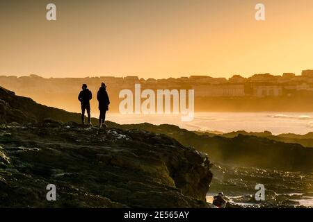 Menschen, die auf Felsen stehen, die von der untergehenden Sonne in Little Fistral in Newquay in Cornwall Silhouette sind. Stockfoto