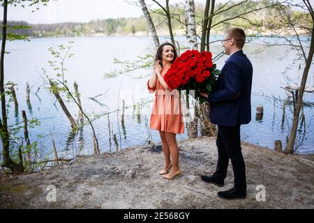 Romantisches Treffen der jungen Leute. Ein Mann in einem Anzug mit einem Strauß roter Rosen gibt dem Mädchen einen Strauß, und sie küssen sich im Wald Stockfoto