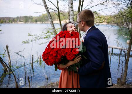 Romantisches Treffen der jungen Leute. Ein Mann in einem Anzug mit einem Strauß roter Rosen gibt dem Mädchen einen Strauß, und sie küssen sich im Wald Stockfoto