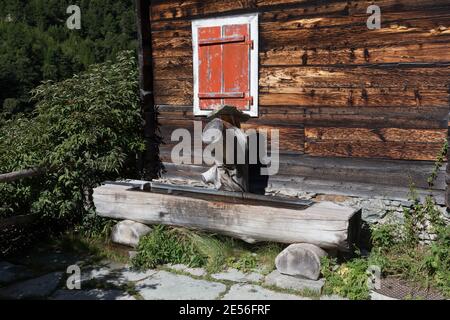 Schweiz Ein traditioneller Holzbrunnen neben einem alten Holzhaus im Alpendorf Zmutt. Stockfoto