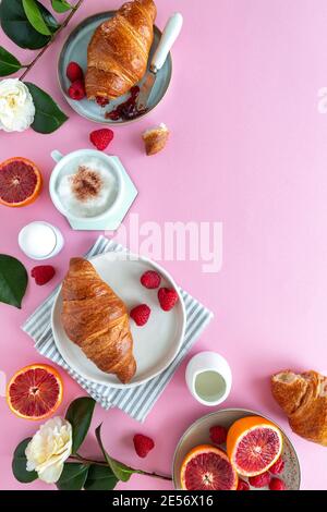 Flaches Lay einer Frühstücksszene mit Croissants, Blutorange, Himbeeren, Blumen und Cappuccino auf rosa Hintergrund Stockfoto