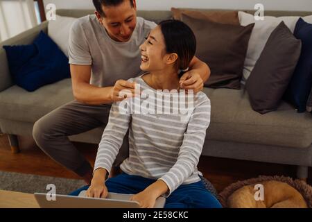 Junger Mann, der seiner Freundin eine Halskette als Geschenk gibt. Asiatische Frau immer überrascht von ihrem Freund mit Heiratsantrag während der Arbeit auf Laptop in modernen Hause Stockfoto