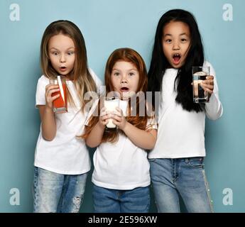 Drei socked Kids Girls Freundinnen in weißen T-Shirts und blauen Jeans stehen mit Gläsern Wasser, Milch und frischem Saft Stockfoto