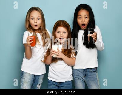 Viel überrascht Kinder Mädchen Freunde in weißen T-Shirts und blaue Jeans stehen mit Gläsern Wasser, Milch und frischen Saft Stockfoto