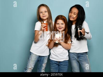 Drei fröhliche Kindermädchen Freundinnen in weißen T-Shirts und blauen Jeans stehen mit ihren Lieblingsgetränken Wasser, Milch und Saft Stockfoto