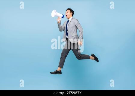Junge asiatische Geschäftsmann springen und schreien auf Megaphon isoliert auf Hellblauer Hintergrund Stockfoto