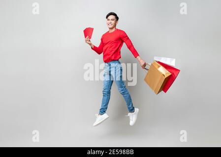 Überrascht asiatischen Mann hält Taschen und roten Umschlag oder Ang PAO springen in isolierten hellgrauen Studio-Hintergrund für Chinesisch New Year Shopping Konzept Stockfoto