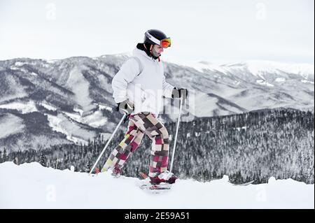 Horizontale Momentaufnahme des jungen Skifahrers in buntem Kostüm, der entlang verschneite Hügel gegen erstaunliche Berge auf Hintergrund läuft, Skitour. Seitenansicht, Kopierbereich, volle Länge Stockfoto