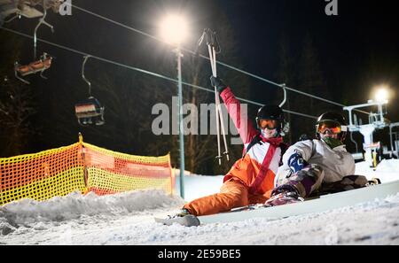 Paar Touristen liegen auf verschneiten Hang unter beleuchteten Sessellift in der Nacht, Ruhe nach aktiven Zeit verbringen. Freunde bei guten Laune, die Spaß im Skigebiet haben Stockfoto